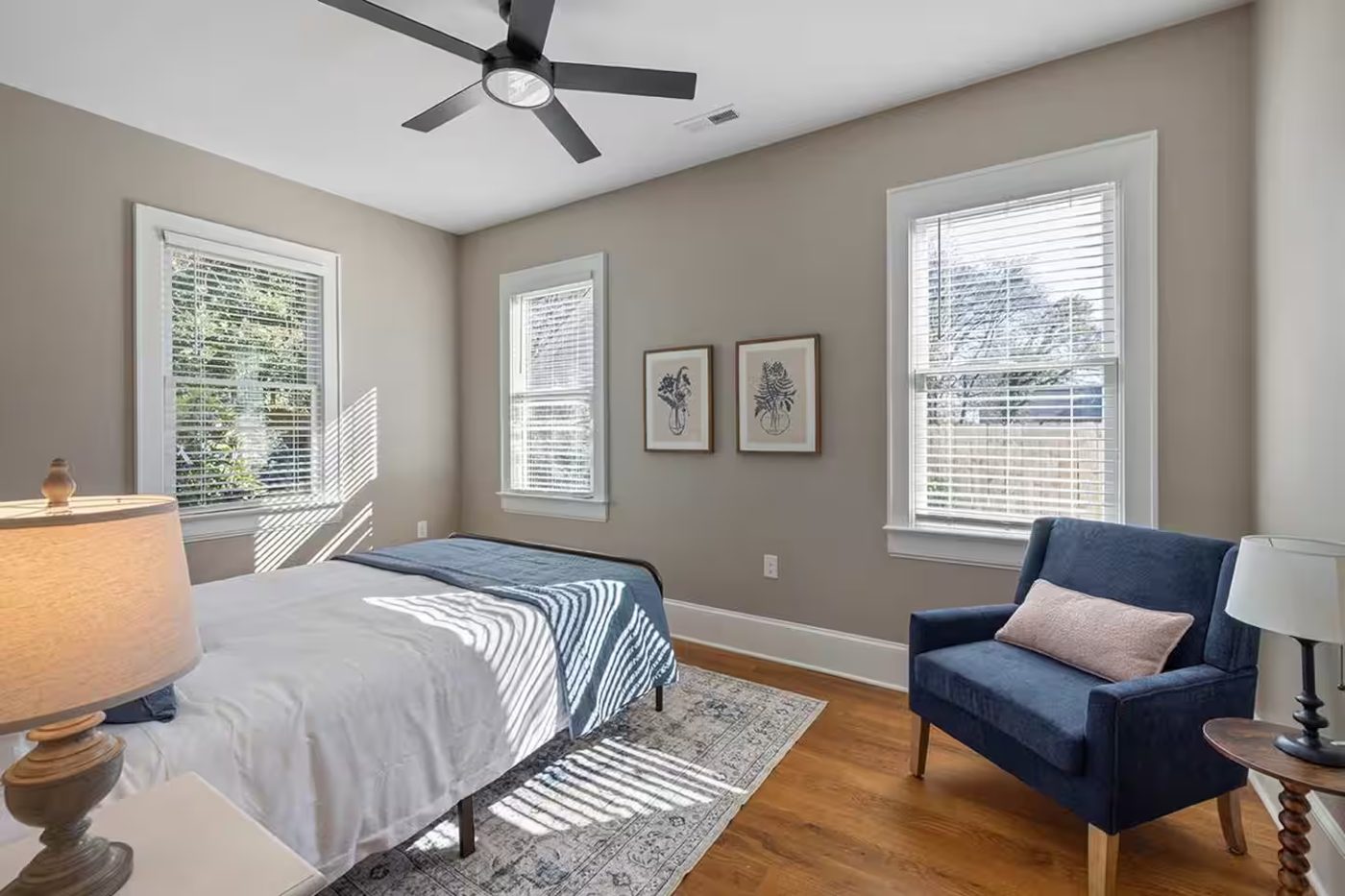 Sunlit bedroom with taupe walls, a navy armchair, a patterned rug, and two framed botanical prints