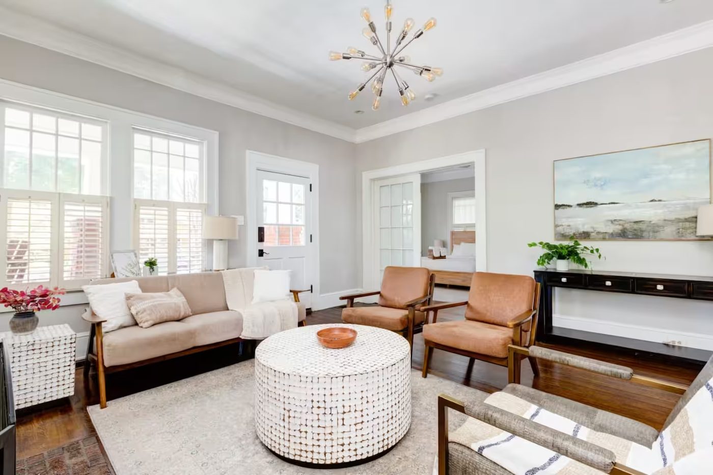 Bright living room with a tan sofa, two leather armchairs, a round textured coffee table, and a starburst chandelier