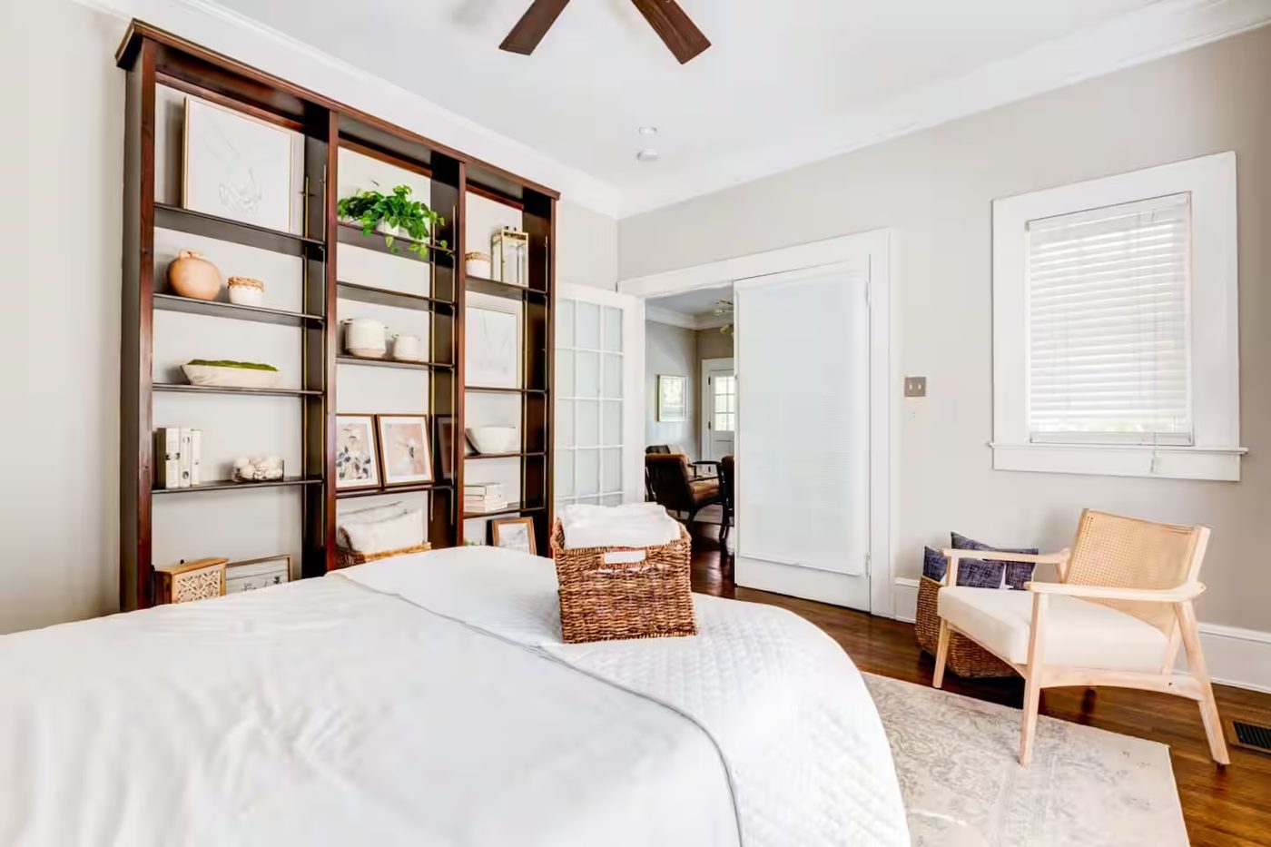 Bedroom with white bedding, a tall dark wood shelving unit, a cane-back chair, and woven baskets