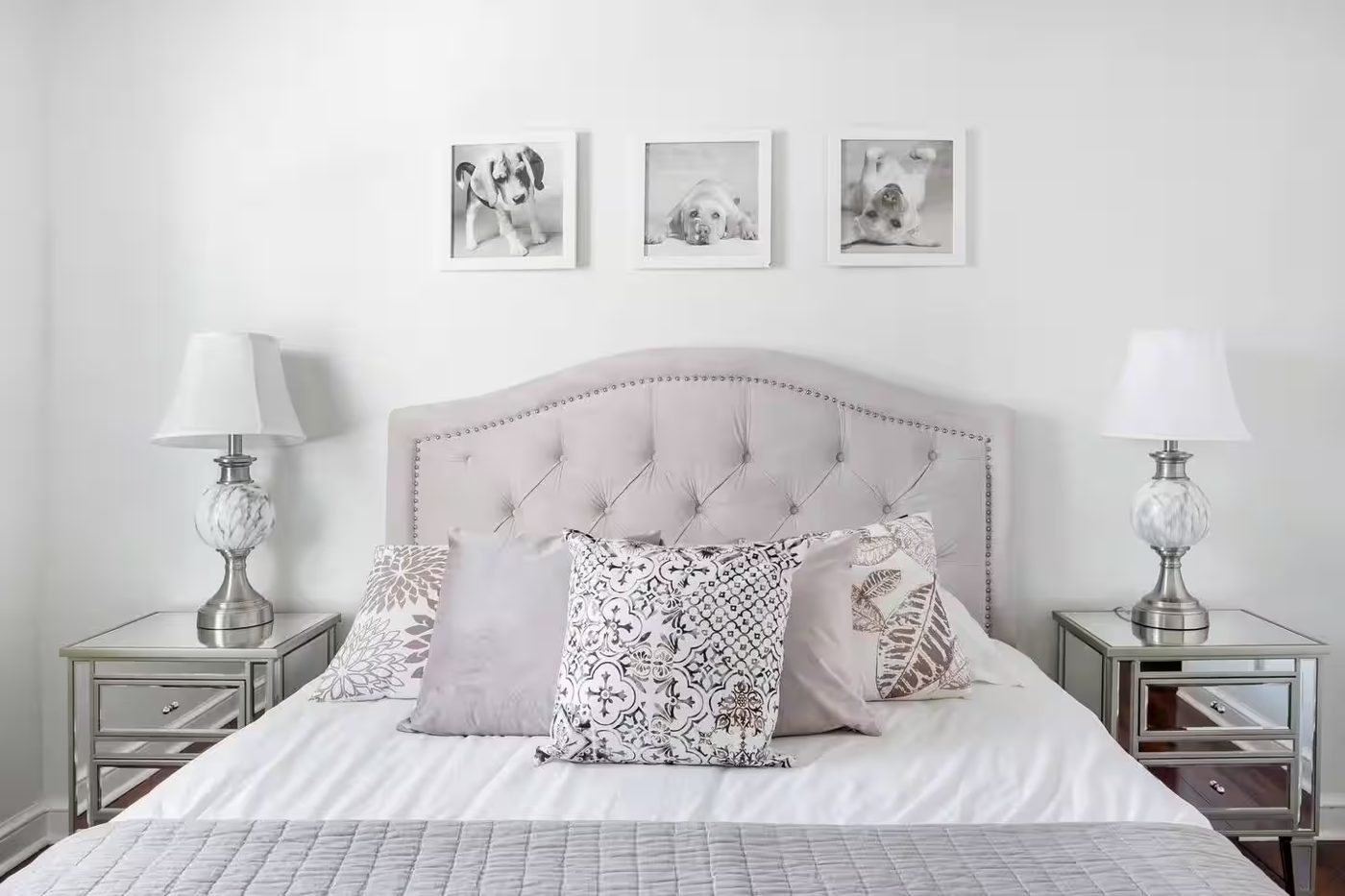 Bedroom with a tufted gray headboard, mirrored nightstands, matching lamps, and three framed dog photos